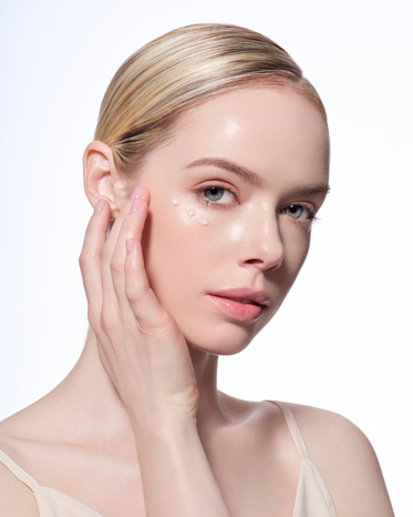 Close-up of a woman with clear skin and blonde hair, has three dots of eye cream under the eyes. She is gently touching her jaw with one hand, showcasing a dewy and radiant complexion.