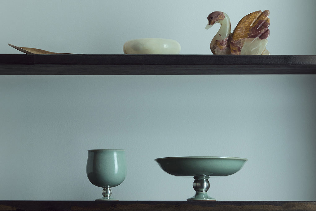 A still-life arrangement of various Korean celadon (청자) ceramic pieces, including a footed dish and tea cups, displayed on a wooden shelf.