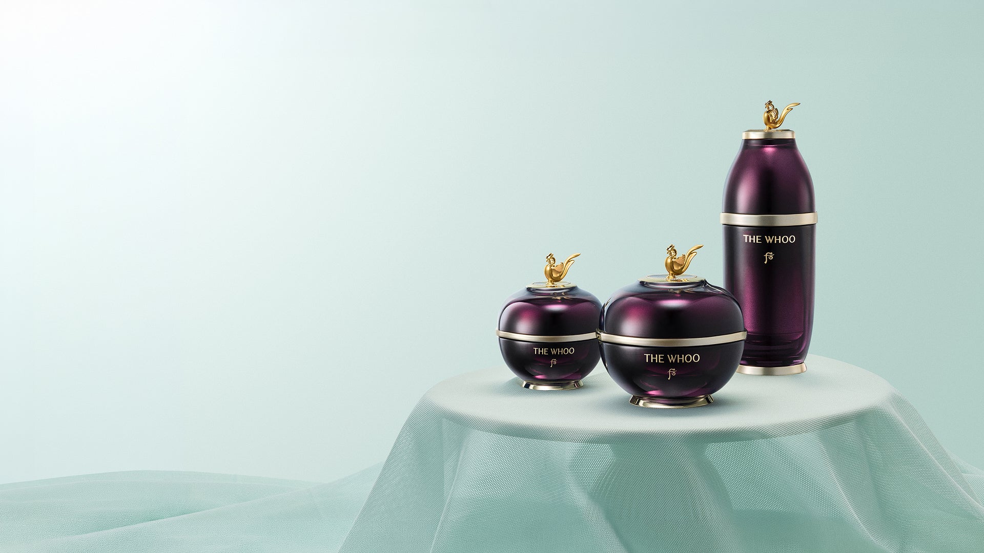 THE WHOO Imperial Youth collection—Cream, Serum, and Eye Cream—elegantly displayed on a soft mint-green silk drape with golden phoenix-inspired accents.