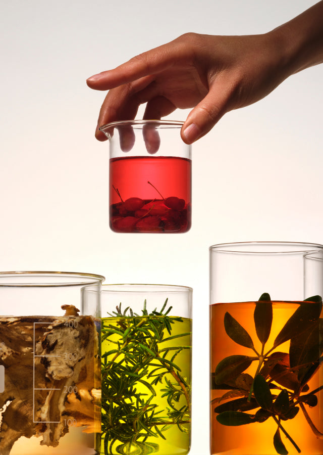 Glass containers filled with natural ingredients and a hand holding a beaker with red extract.