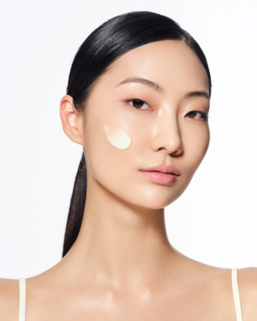 A women with sleek, dark hair styled in a low ponytail, has a small amount of cream applied to her cheek. She is showcasing a smooth neck and shoulders, wearing a thin-strap top against a plain white background.