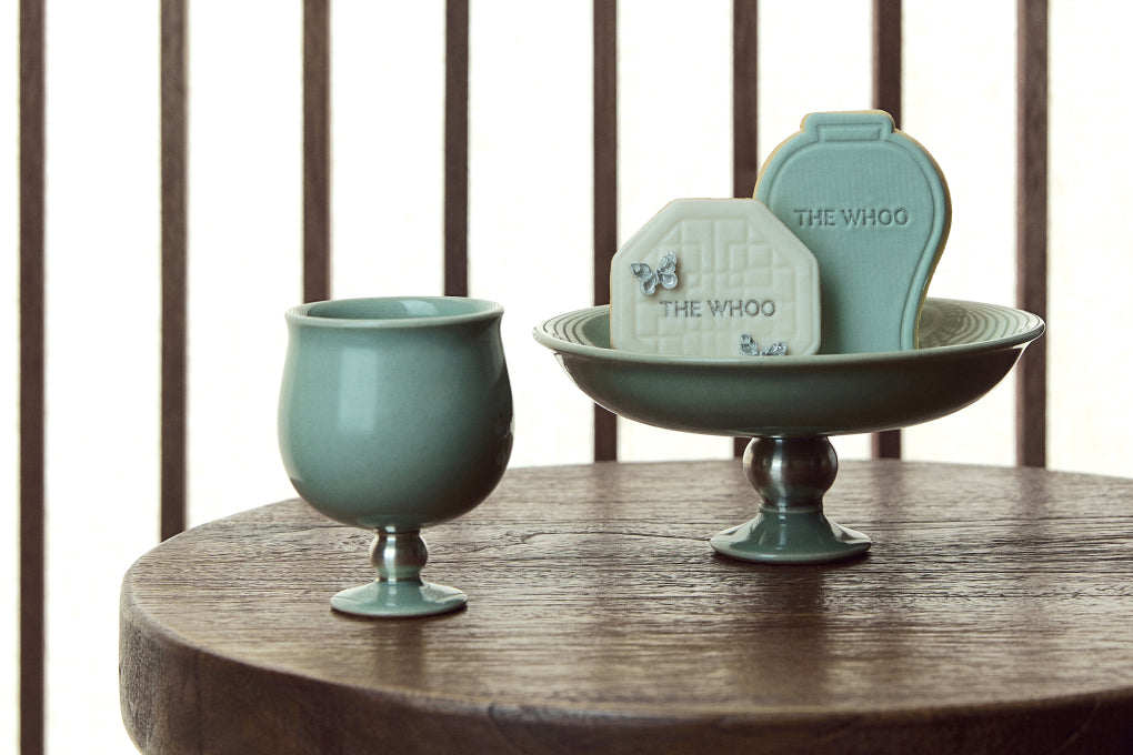 A curated display of the THE WHOO x YERAMIC collaboration, featuring a celadon vase, a footed tray, and decorative ceramic tags on a dark wooden surface.